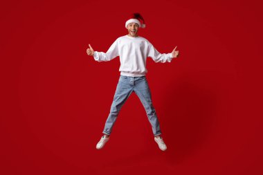 A cheerful individual wearing a Santa hat jumps with excitement, showcasing a thumbs-up gesture. The bright red background enhances the holiday spirit, capturing the joy of the season.