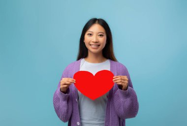 Göğsüne yakın büyük kırmızı bir kalp kesiği tutan uzun siyah saçlı bir kadın. Açık mavi arka plan neşeli bir atmosfer yaratır. Açık renk bir gömleğin üzerine hoş mor bir hırka giyiyor..