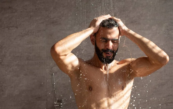 Genç Orta Doğulu adam ferahlatıcı bir duşun altında duruyor, gözleri kapalı ve elleri saçında. Modern bir banyo ortamında su çağlayarak onu aşağı indiriyor ve rahatlatıcı bir atmosfer yaratıyor..