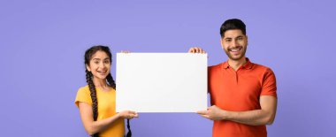 İki gülümseyen arkadaş birlikte durup boş beyaz bir işaret tutuyorlar. Yumuşak mor bir arka planı olan canlı bir yerdeler. Atmosfer eğlenceli ve davetkar. Yaratıcı mesajlar için mükemmel..