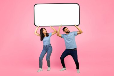 Genç bir çift, büyük boş bir tabela tutarken gülümsüyor. Parlak pembe bir arka planın önündeler ve rahat giyiniyorlar, neşeli ve neşeli görünüyorlar..