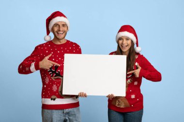 Noel süveteri ve Noel Baba şapkaları giyen neşeli bir çift gülümseyerek ve boş bir tabela tutarak yan yana duruyorlar. Açık mavi bir arkaplanın önünde neşeli bir hava yaratıyorlar..