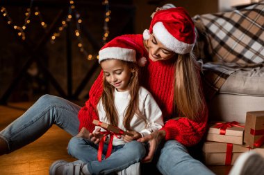 Noel için dekore edilmiş sıcak bir oturma odasında, festival şapkalı bir anne ve kız hediyeleri birlikte açmaktan keyif alıyorlar. Atmosfer neşe ve aşkla dolu..