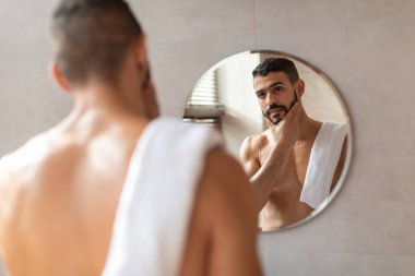 Yakışıklı genç bir Arap banyo aynasının önünde duruyor. Görünüşüne hayran kalarak çenesine dokunuyor..