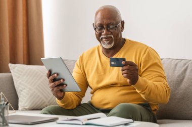 Son sınıf öğrencisi rahat bir şekilde kanepede oturur, tabletine bakar ve elinde bir kredi kartı tutar, rahatlatıcı bir öğleden sonra internetten alışveriş yapar..