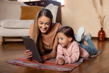 A mother and her daughter are lying on a colorful rug, engaged in an activity on a tablet. They are smiling and clearly enjoying their time together in a warm living room.