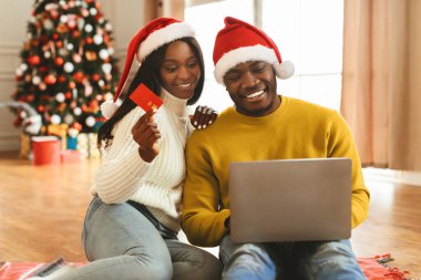 İki kişi dizüstü bilgisayara bakarken yerde oturuyor. Birinin kredi kartı var. Noel şapkası takarlar ve rahat bir odada tatil süslemeleriyle çevrilidirler..