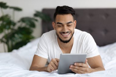 Orta Doğulu genç bir adam rahatça yatakta yatarken gülümsüyor, dijital tabletini karıştırıyor. Çevrimiçi içerikle ilgileniyor, muhtemelen alışveriş yapıyor veya çeşitli ilgi alanlarını araştırıyor..