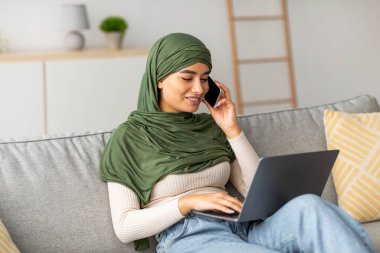 Genç bir Arap kadın kanepede oturuyor, telefonla konuşurken dizüstü bilgisayar kullanıyor. Rahat bir ortamda evden bağımsız işini yönetirken odaklanmış ve pozitif görünüyor..