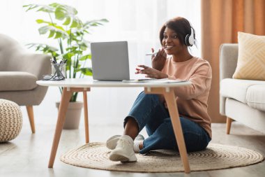 Bir kadın, küçük bir masada dizüstü bilgisayar kullanırken gülümseyerek yerde rahatça oturuyor. Kulaklık takıyor. Etrafı bitkilerle ve yumuşak mobilyalarla çevrili..