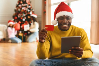 Kırmızı şapkalı bir adam alışveriş için tablet kullanırken yerde oturup gülümsüyor. Arka planda, aile üyeleri süslenmiş bir Noel ağacının etrafında toplanmış, şenliğin tadını çıkarıyorlar..
