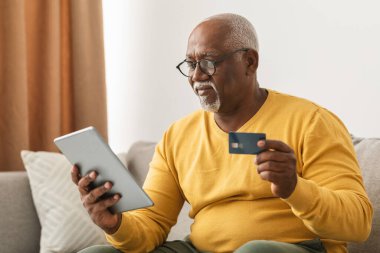 Yaşlı bir adam kanepede rahatça oturup elinde kredi kartıyla online alışveriş yapıyor. Sıcak oturma odası, rahat bir atmosfer yaratıyor..
