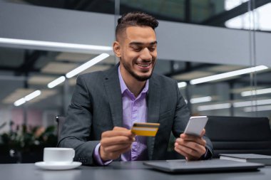 Bir adam gülümseyen ve akıllı telefonuna bakan bir masada oturuyor. Elinde bir kredi kartı tutuyor. Modern bir ofis ortamında onun yanına bir fincan kahve konur..