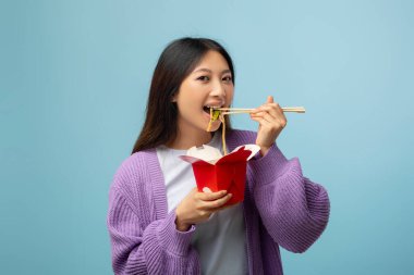 Genç bir kadın elinde eriştelerle dolu kırmızı bir paket yemek kutusu tutuyor. Yemek çubuklarıyla gülümsüyor. Rahat, mor bir süveter giyiyor, canlı bir atmosfer yaratıyor..