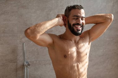 Yakışıklı genç bir Arap erkek model modern bir banyoda sıcak bir duş alırken sevinçle gülümsüyor. Kafasını şampuanla yıkıyor. Düşen su damlalarının altında..
