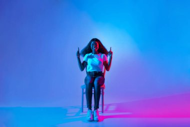 Bir kadın, mavi ve pembe ışıkların parlak arka planına karşı bir sandalyede oturuyor. Gülümser ve parmaklarını şakayla kaldırır. Yaratıcı stüdyo ortamında neşeli bir anı yakalar..