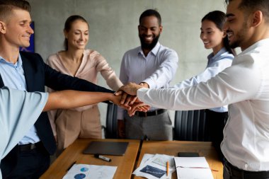 Bir şirket ofisindeki çeşitli meslektaşlar el ele tutuşarak başarılarını kutlamak için bir araya gelirler. Bu an, bir toplantı sırasında ekip üyeleri arasındaki birlik ve işbirliğini gösteriyor.