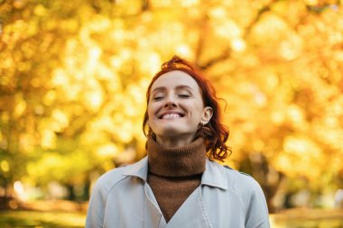 Kızıl saçlı bir kadın sonbaharda bir parkta dururken neşeyle gülümser. Arka plan sıcak güneş ışığı altında parlak sarı yapraklarla dolu, neşeli bir atmosfer yaratıyor..