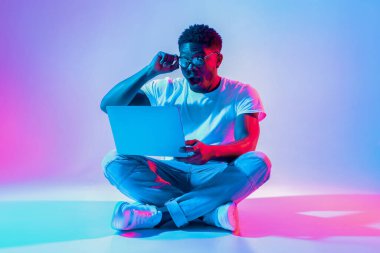 Genç bir adam dizüstü bilgisayar kullanırken bağdaş kurarak yerde oturuyor. Stüdyoda canlı mavi ve pembe ışıklar var, şık bir atmosfer yaratıyor. Ekrana odaklanmış durumda..