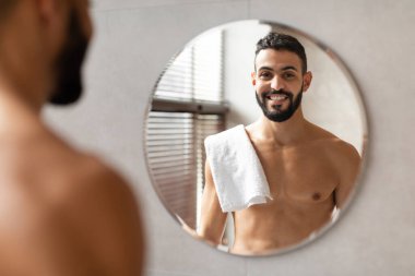 Sakallı genç bir Orta Doğulu aynadaki yansımasına gülümsüyor. Omzunda beyaz bir havluyla şık bir banyoda duruyor. Kendine güveni ve pozitifliği var..