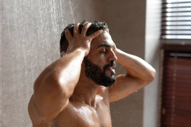 Yakışıklı genç Arap erkek modern bir duşta, gözleri kapalı, saçına şampuan sürüyor. İçerideki dingin ortam, erkek güzellik alışkanlığını geliştiriyor, özel ilgi gösteriyor..