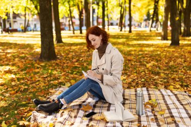 Bir kadın sonbaharda bir parkta bir battaniyeye oturur. O, parlak sarı ve turuncu yapraklarla çevrili açık renk bir ceket giyerek defterine yazıyor..