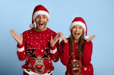 Neşeli bir çift birlikte poz veriyor, rengeyiği tasarımları ve Noel Baba şapkalarıyla bezenmiş kırmızı kazaklar giyiyorlar. Tatil sezonunun neşeli atmosferini kutluyorlar..