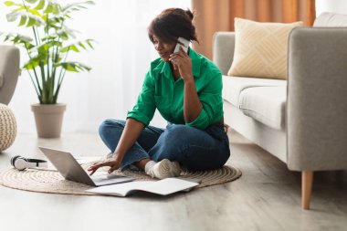 Bir kadın dizüstü bilgisayarı ve defteriyle yerde oturmuş telefonla konuşuyor. Oturma odasında. Pencereden içeri doğal ışık giriyor..