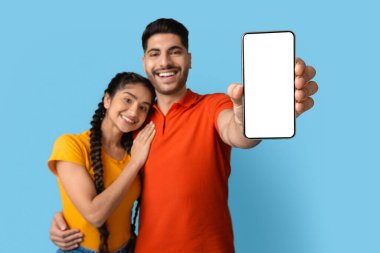 Genç bir çift birbirine yakın duruyor, kameraya doğru boş bir akıllı telefon tutarken geniş bir gülümseme sergiliyorlar. Neşeli ifadeleri, canlı mavi bir zemin karşısında aşkı ve mutluluğu ifade eder..