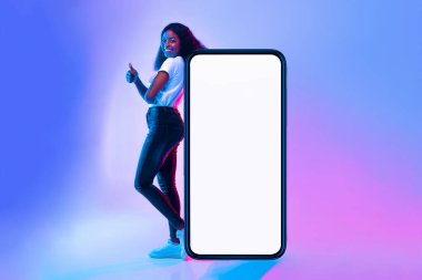 Genç bir kadın boş ekranlı büyük bir akıllı telefonun yanında duruyor, gülümseyerek başparmağını kaldırıyor. Renkli arka plan mavi ve pembe tonlarını içeriyor, canlı bir atmosfer yaratıyor..