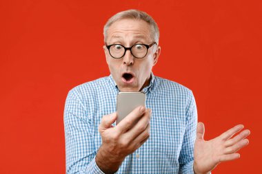 Gözlüklü yaşlı bir adam parlak kırmızı bir arkaplan önünde bir akıllı telefonu tutarken şaşırmış görünüyor. Yüz ifadesi ekranda gördüklerine olan inancını ve şaşkınlığını gösteriyor..