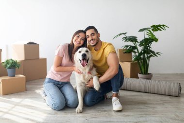 Neşeli bir çift yerde köpekleriyle oturuyor, gülümsüyor ve birbirlerine eşlik ediyorlar. Arka planda taşınan kutular yeni bir eve taşındıklarını gösteriyor..