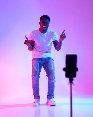 Genç bir Afrikalı Amerikalı erkek etkili akıllı telefon kamerasıyla neon ışıklı canlı bir ortamda neşeyle bağlanıyor. Çevrimiçi takipçileri için eğlenceli video içeriği yaratıyor..