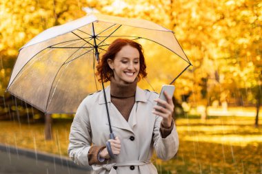 Sonbahar parkında şeffaf bir şemsiye altında akıllı telefonunu kontrol ederken kızıl saçlı bir kadın gülümsüyor. Ağaçlar altın yapraklarla doludur. Sıcak ve canlı bir sahne yaratırlar..