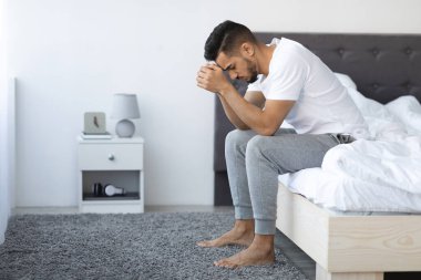 Genç bir Arap, basit bir yatak odasında yatağının kenarında oturuyor. Kafasını elinde tutarak sıkıntılı görünüyor. Atmosfer umutsuzluk ve ilgisizliği yansıtıyor..