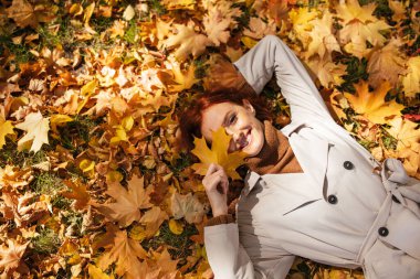 Bir kadın, canlı sonbahar yapraklarıyla kaplı yerde dinlenir. O açık renkli bir ceket giyer ve bir yaprak ile şakayla yüzünü örterek gülümser. Sahne sonbaharın neşesini yakalar..
