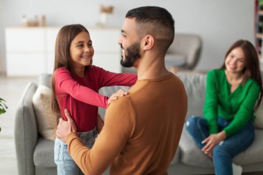 Küçük bir kız, babalar gününü kutlamak için sıcak bir oturma odasında babasını kucaklıyor. Mutlu bir anne kanepede oturup sıcak bir aile anı yaratırken gülümser..