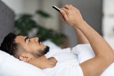 Genç bir Arap adam internette gezinmek için akıllı telefonunu kullanarak yatakta yatıyor. Rahat bir yatak odasında, modern dekorlar sergiliyor. Nişanlı ifadesine odaklanmış durumda..