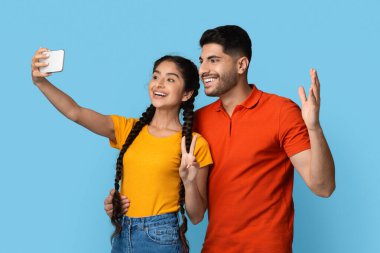 Genç bir erkek ve kadın selfie çekerken geniş bir gülümseme sergilerler. Erkek el sallarken, kadın barış işareti gösterirken mutlu poz veriyorlar. Arkaplan parlak mavi.