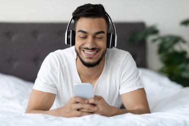 Yakışıklı bir Orta Doğulu genç adam kablosuz kulaklık takarak yatakta rahatça yatıyor. Akıllı telefonuyla mesajlaşırken gülümsüyor, rahat yatak odası atmosferinin tadını çıkarıyor..