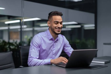 Ofis ortamında bir kişi dizüstü bilgisayarla çalışıyor. Çalışma alanı modern mobilyalara ve parlak ışıklandırmaya sahiptir. Adam bilgisayarı kullanırken meşgul ve mutlu görünüyor..