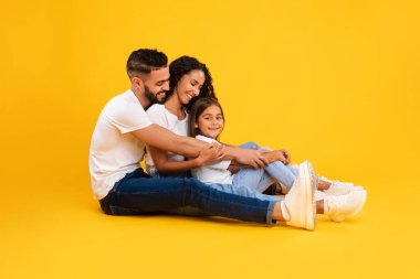 Üç kişilik neşeli bir aile, canlı sarı bir zemin üzerinde oturuyor. Ebeveynler, mutlu bir şekilde gülümseyen kızlarıyla şakalaşıyorlar..