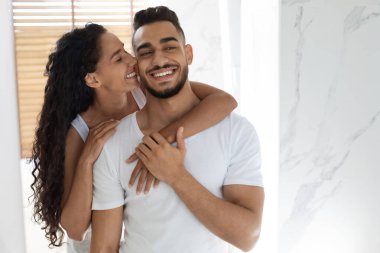 Mutlu bir çift, iyi ışıklandırılmış bir odada gülümseyerek ve sevgisini paylaşarak duruyor. Kadın şakacı bir şekilde erkeği yanağından öper, sıcak ve sevgi dolu bir atmosfer yaratır..