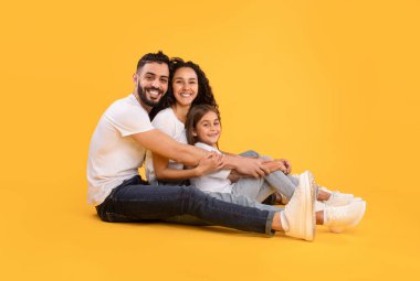 Mutlu bir aile, canlı sarı bir arka planda yan yana oturur. Bir baba, anne ve kız mutlu bir anı paylaşırken gülümserler, bağlarını ve sevgilerini vurgularlar..