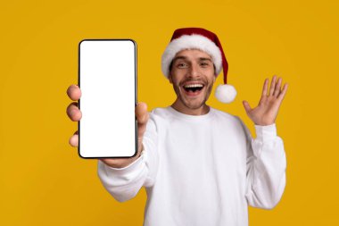 Noel Baba şapkası takan genç bir adam gülümsüyor ve elinde boş bir ekranla akıllı telefon tutuyor. Parlak sarı bir arka planda dururken neşeli ve neşeli görünüyor..