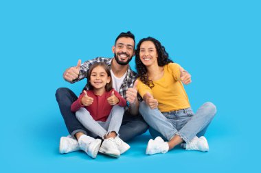 Üç kişilik neşeli bir aile yerde oturuyor. Hepsi başparmağını kaldırır ve mutlu bir ifade takınırlar. Arka plan parlak mavi, neşeli bir atmosfer yaratıyor..