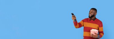 Kocaman gülümseyen bir adam bir elinde patlamış mısır, diğer elinde uzaktan kumanda tutuyor. Mobil cihazında mavi arka planda eğlenceli bir şey izlerken heyecanlı görünüyor..