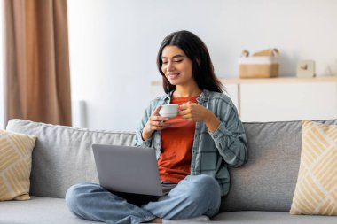 Young Arab woman sits cross legged on a couch, focused on her laptop. She enjoys a cup of coffee while studying remotely and engaged in online work in a cozy living room.