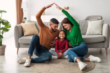 Sıcak bir oturma odasında gülümseyen Arap ebeveynler mutlu kızlarını kucaklayan bir çatı oluşturuyorlar. Aile bir halının üzerinde birlikte oturur, sevgi ve mutluluk anlarını paylaşır..