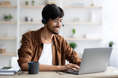 Genç bir adam, rahat bir ofiste çalışarak verimli bir günün tadını çıkarıyor. Kulaklık takıyor ve bir dizüstü bilgisayarı açık gülümsüyor, görevleriyle meşgul..